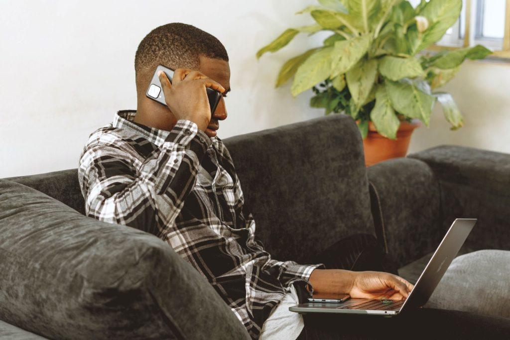 Young man multitasking with phone and laptop on a sofa, working remotely from home.
