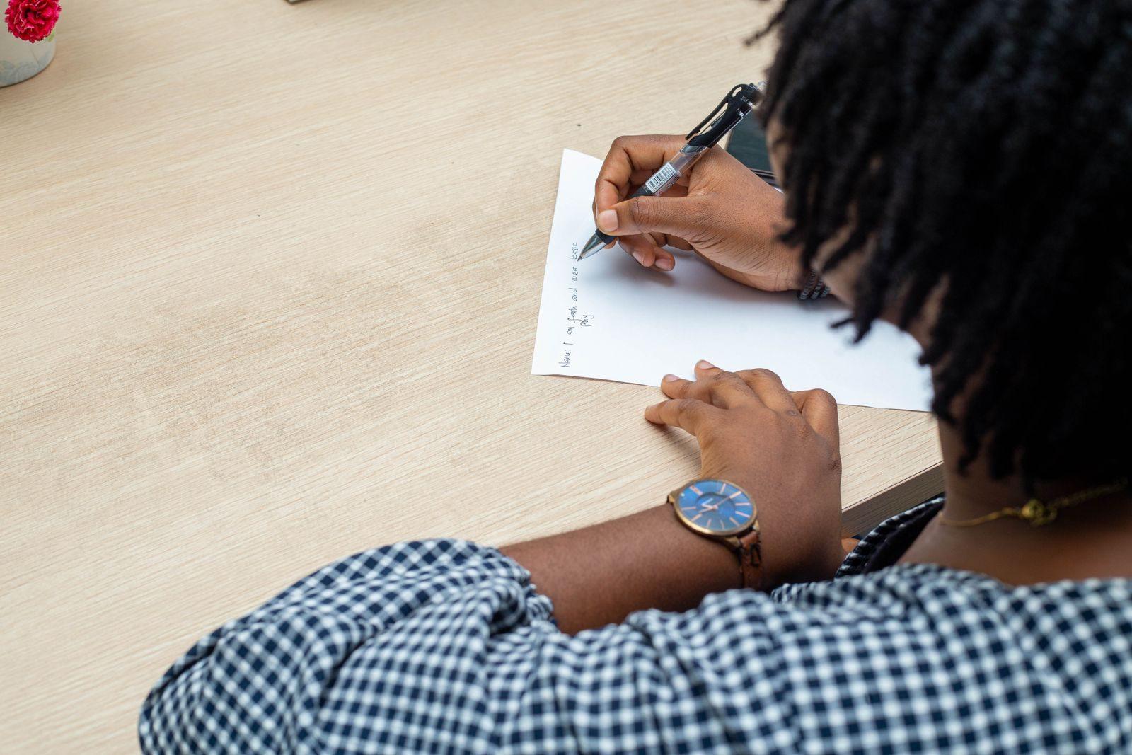 An individual writing notes on paper at a wooden desk in Lagos, Nigeria, wearing a wristwatch.