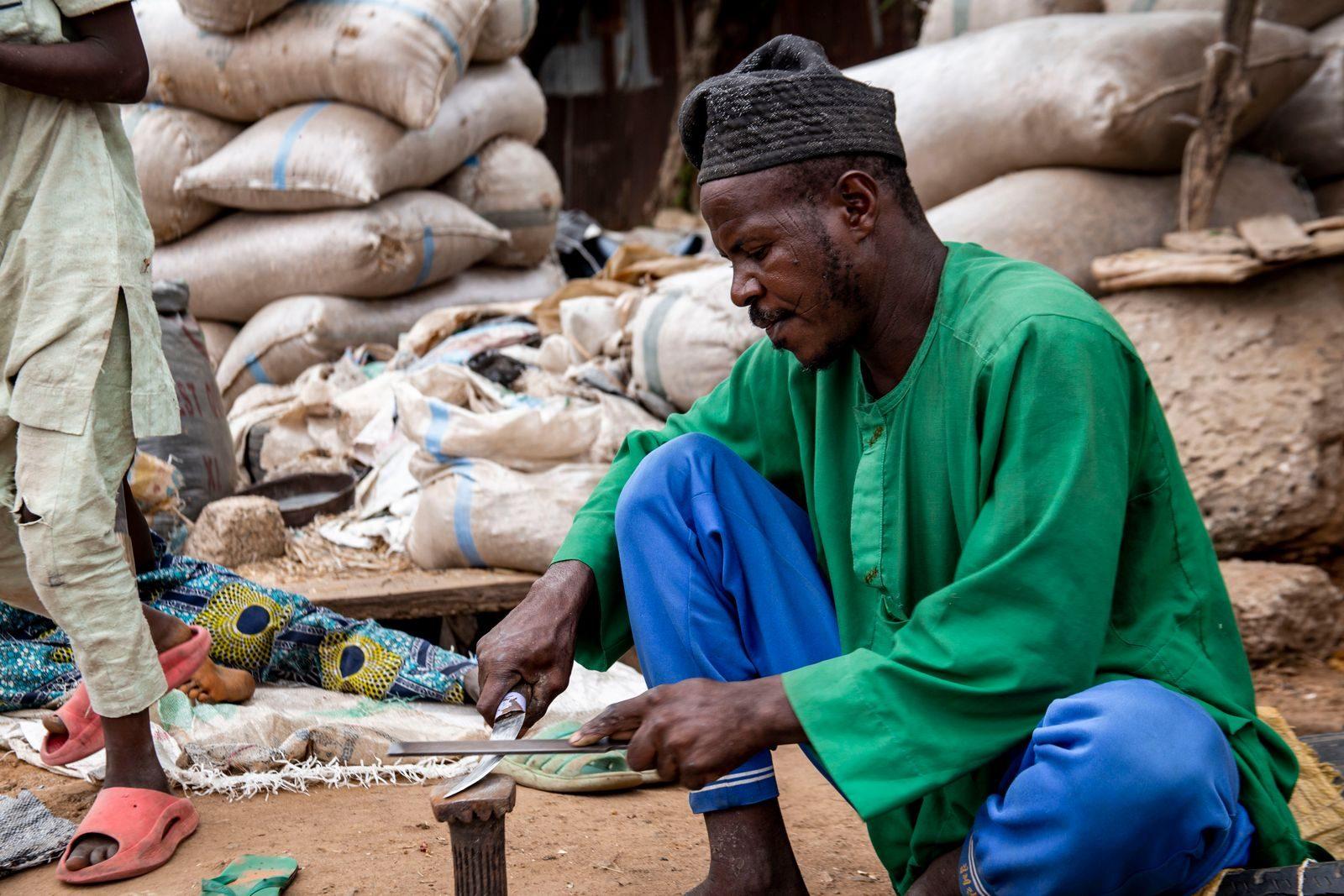 A Nigerian man skillfully crafts in a bustling market surrounded by sacks in vibrant attire.