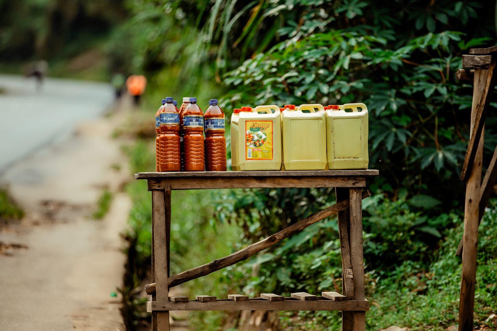 Rural roadside stall featuring bottles and jerrycans of oil and juice with a lush green backdrop.