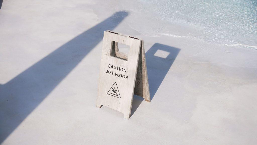 A caution wet floor sign placed near a poolside in bright daylight, casting shadows on the ground.