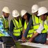 A diverse team of five engineers in safety gear examines construction plans indoors.