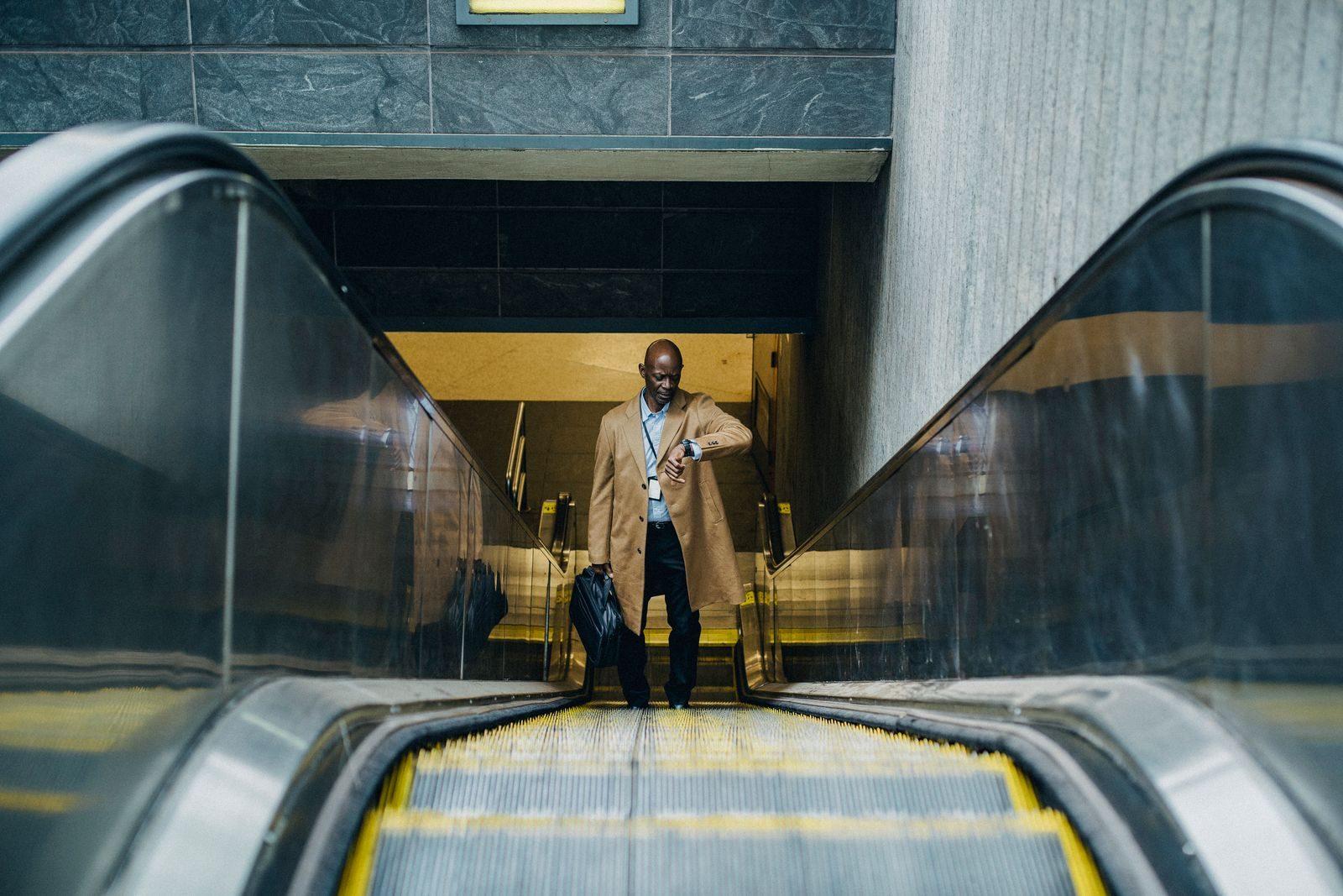High angle full length pensive African American businessman in formal clothes with laptop case riding escalator and checking time on wristwatch while leaving metro station