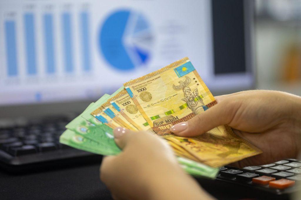 Hands holding Kazakhstan tenge bills with financial chart and graphs on computer screen.
