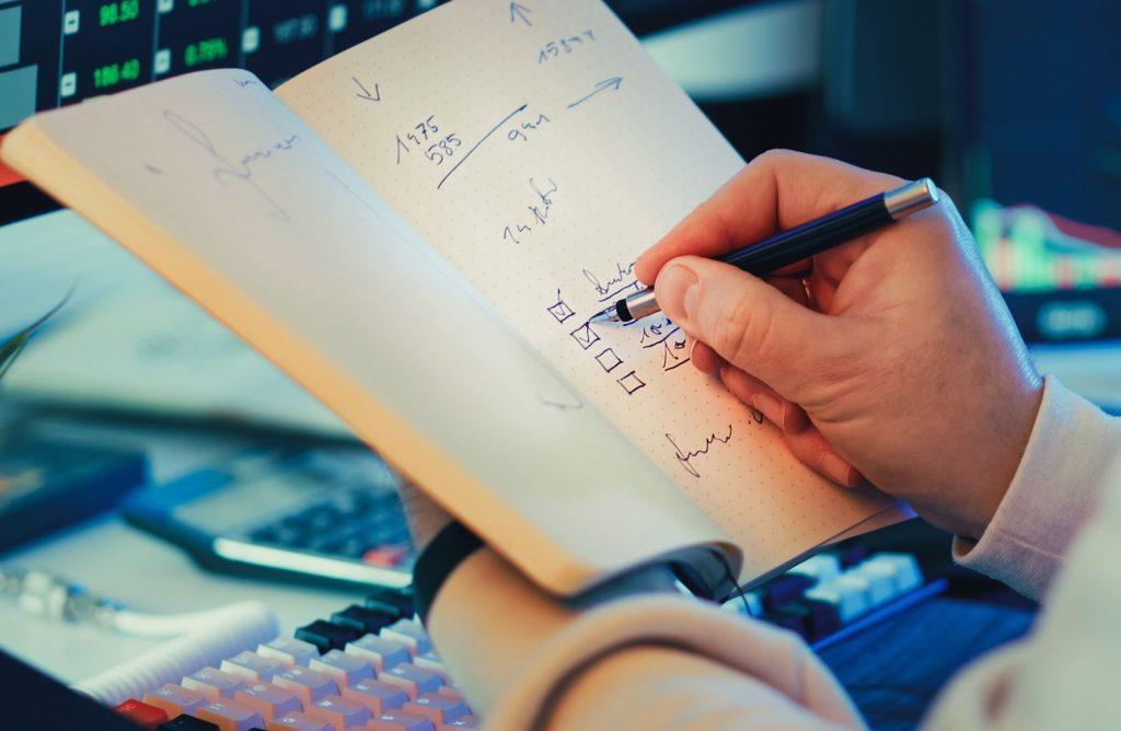Photo by Jakub Zerdzicki A hand writing in a notebook during stock market analysis on a desk.
