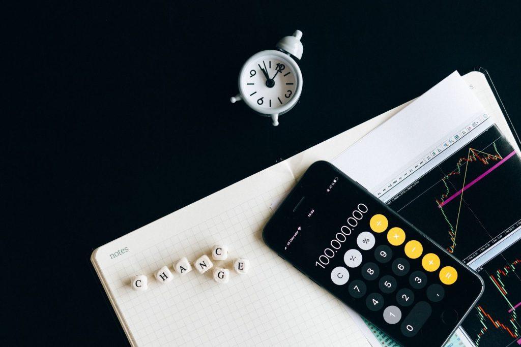 Photo by Nataliya Vaitkevich Notebook with an iPhone displaying 100,000,000, desk clock and financial charts suggesting market analysis.