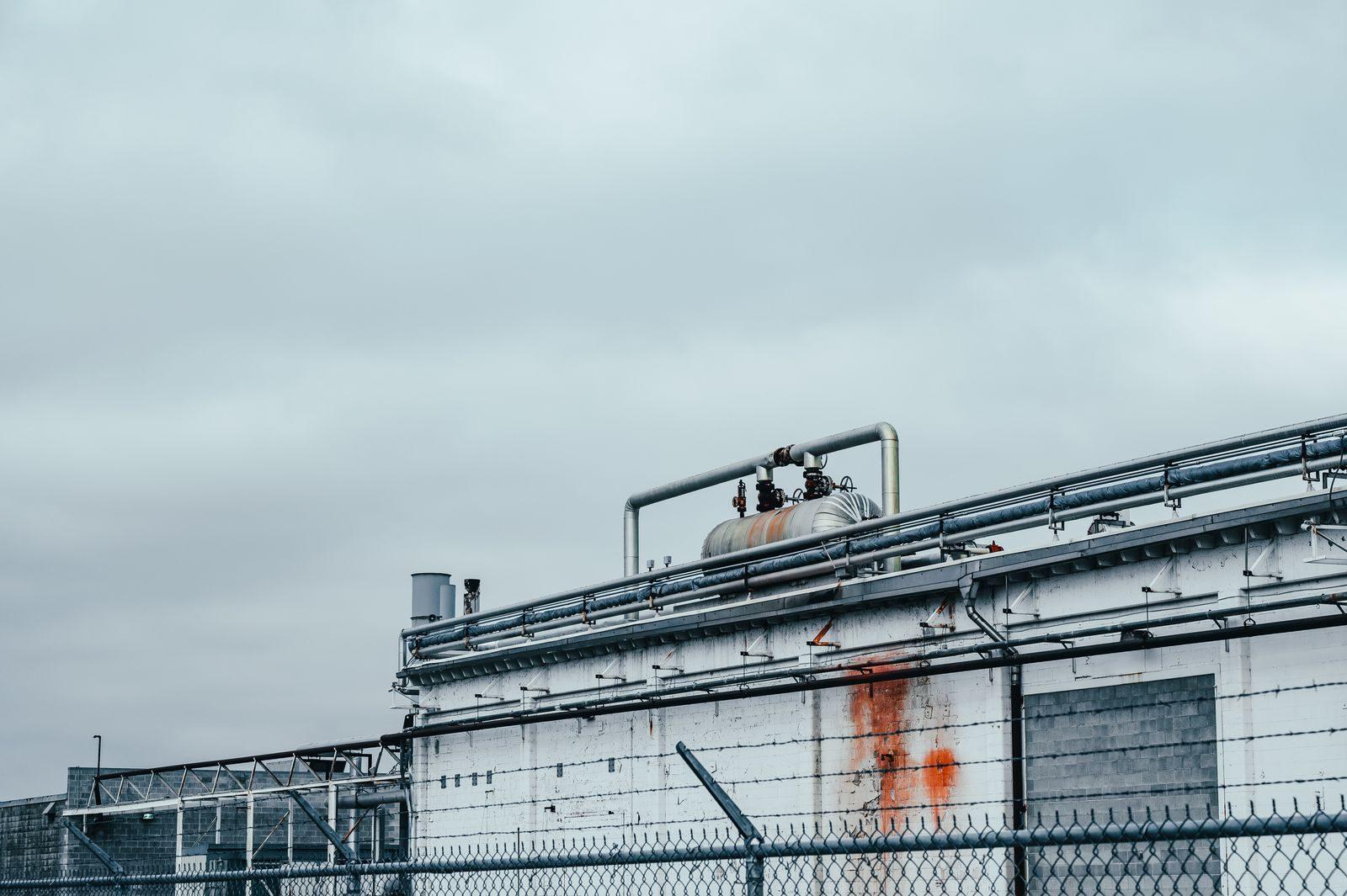 Shabby wall of factory with metal pipes and special equipment located near fence in industrial district of town against cloudy sky