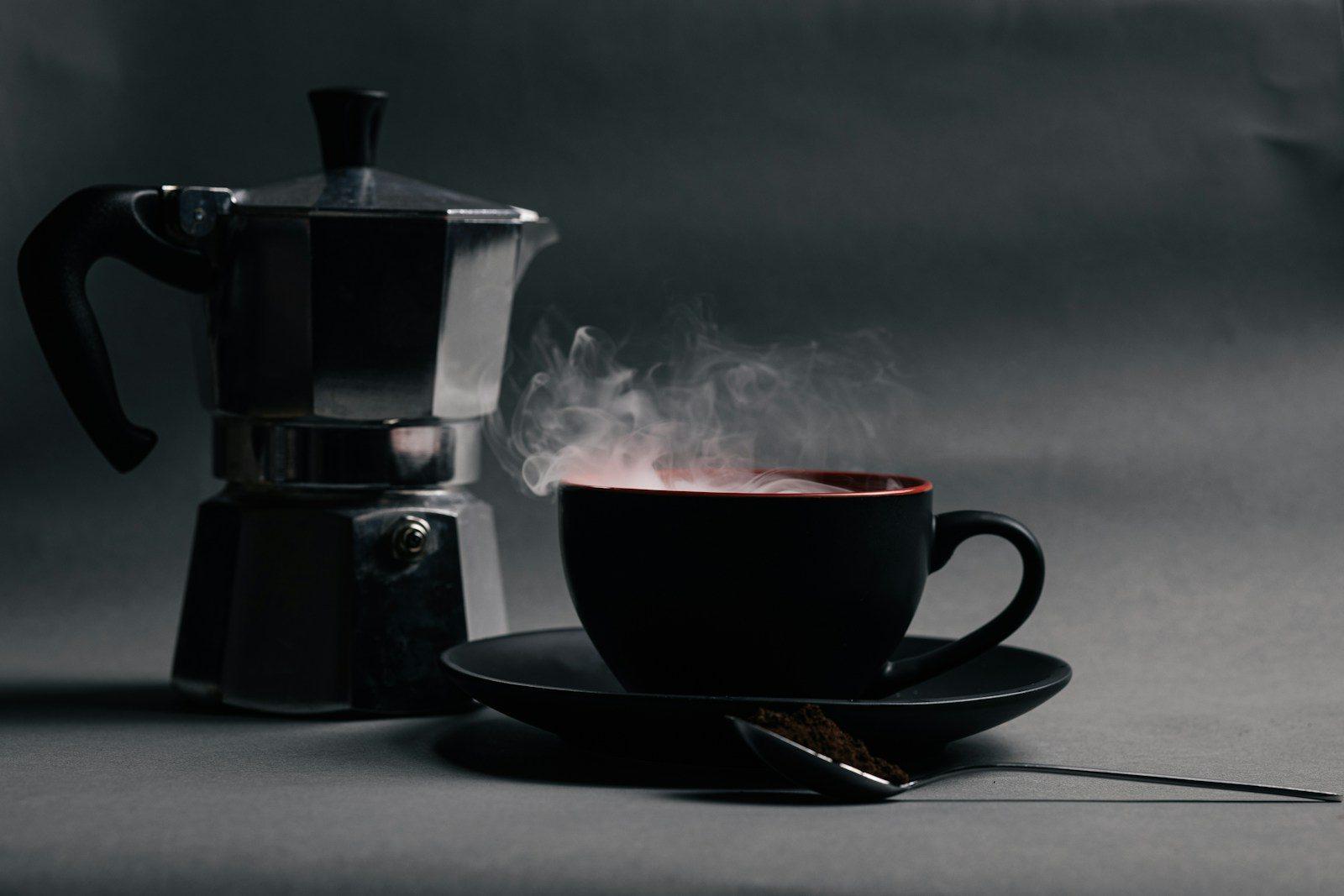 A steaming black coffee cup beside a metal moka pot on a dark grey surface
